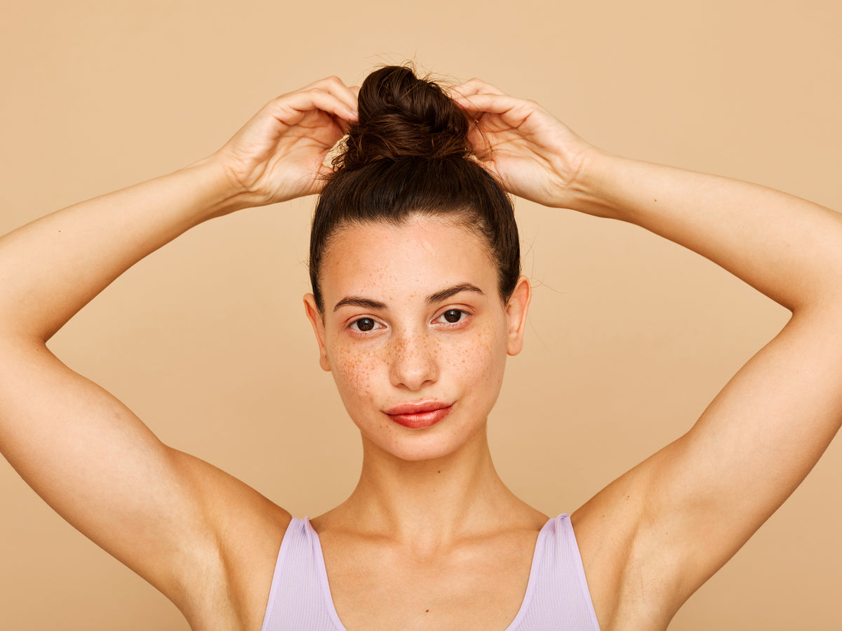 Young woman with freckles and a top bun against a beige background. She's wearing a lavender tank top and looks confident and serene.