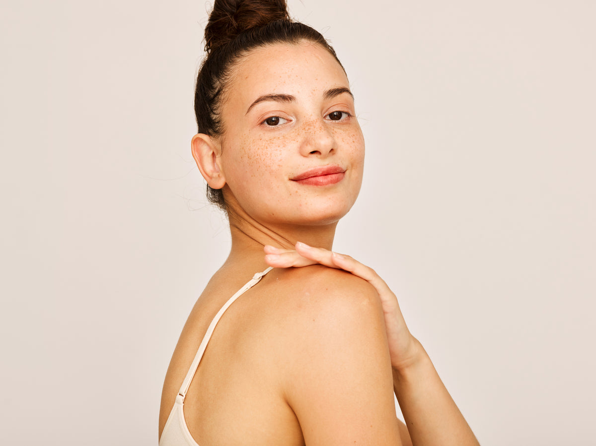 A woman with a bun hairstyle and freckles gently touches her shoulder. She wears a beige top and smiles slightly against a neutral background.