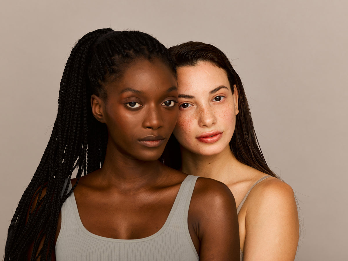 Two women stand closely against a neutral background. One has braided hair and wears a gray top, the other has straight hair with freckles, both with serene expressions.