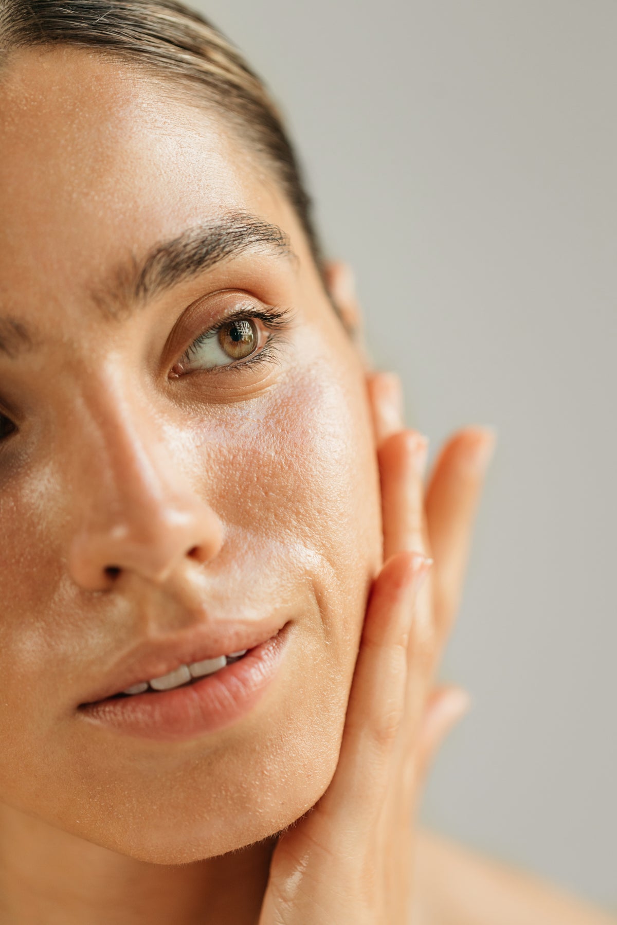 Close-up of a woman gently touching her cheek, showcasing smooth, glowing skin with light makeup. The expression is serene and thoughtful.