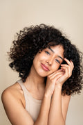 A woman with curly hair and a gentle smile poses against a beige background, resting her cheek against here hands.