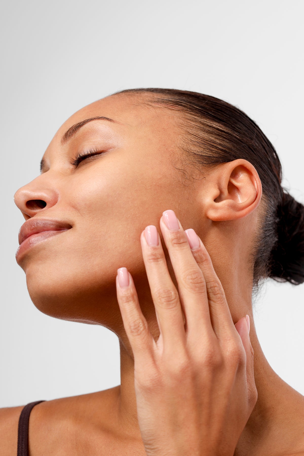 A woman with eyes closed touches her smooth cheek with manicured fingers. Her hair is neatly pulled back, set against a soft grey background.